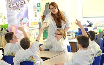 Mad Scientist passing stack of activity cups to student while kids in classroom look on