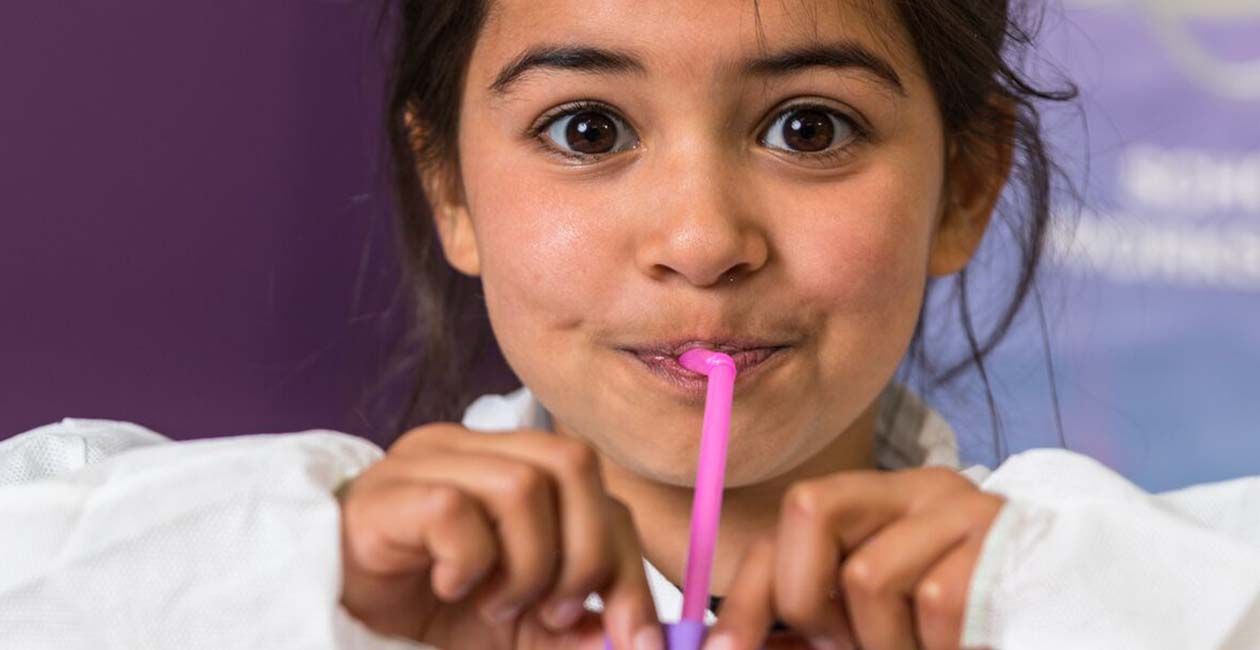 Girl in lab coat blowing up purple balloon using a straw